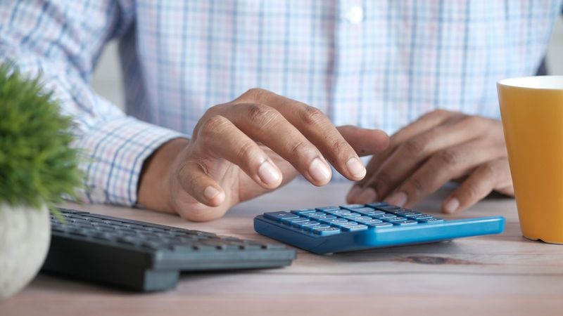 A person's hands typing figures into a blue calculator beside a keyboard