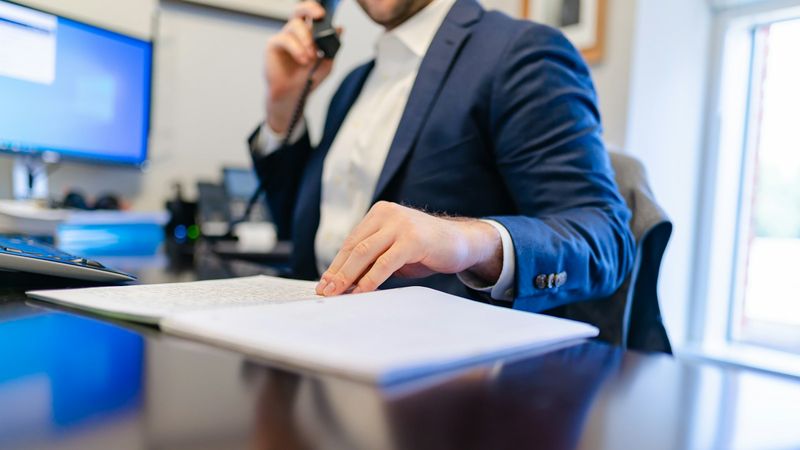 An office worker on the desk phone keeps a hand on a page of handwritten notes, a computer screen beside him