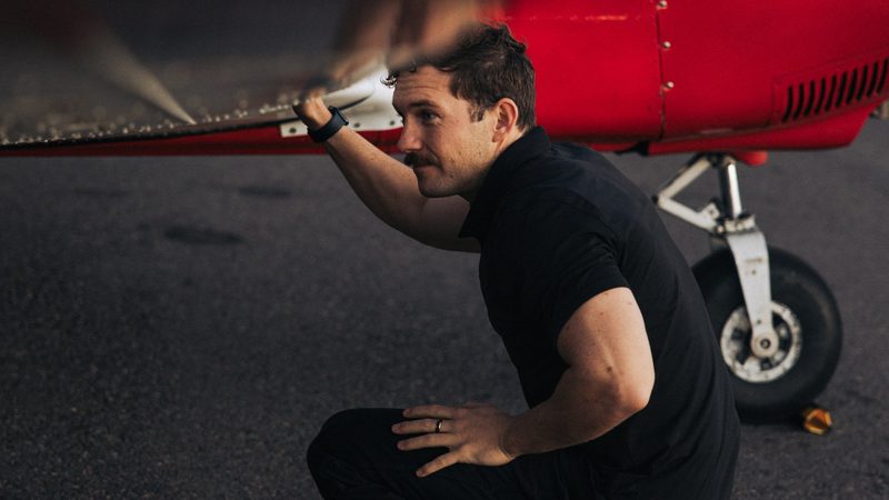 A mechanic crouches under the wing of a small red airplane and checks its underside