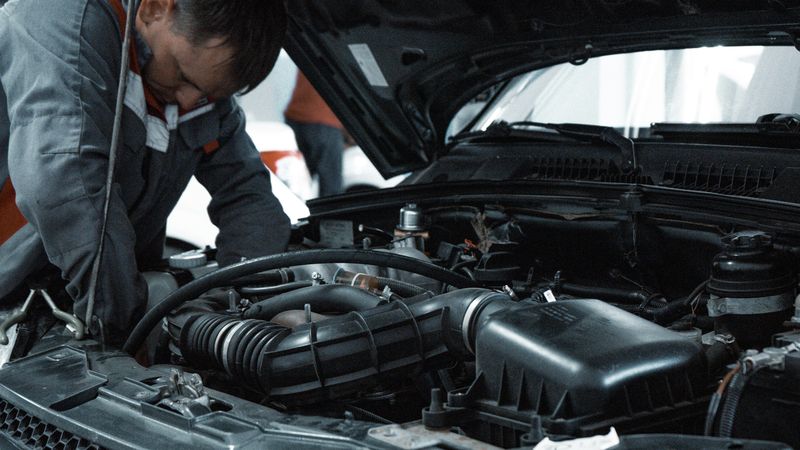 An auto mechanic in a grey work jacket leans into the open engine bay of a car