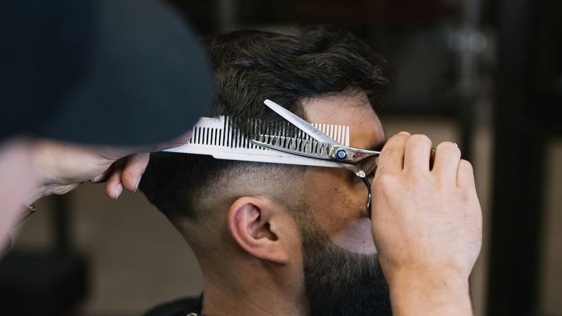 A barber trims a bearded client's hair with a comb and thinning shears