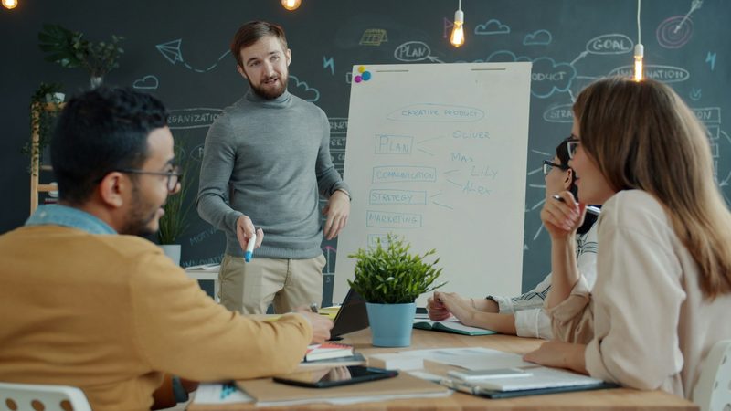 A man with a marker talks to three colleagues at a table in front of a flip chart that lists plan, strategy and marketing