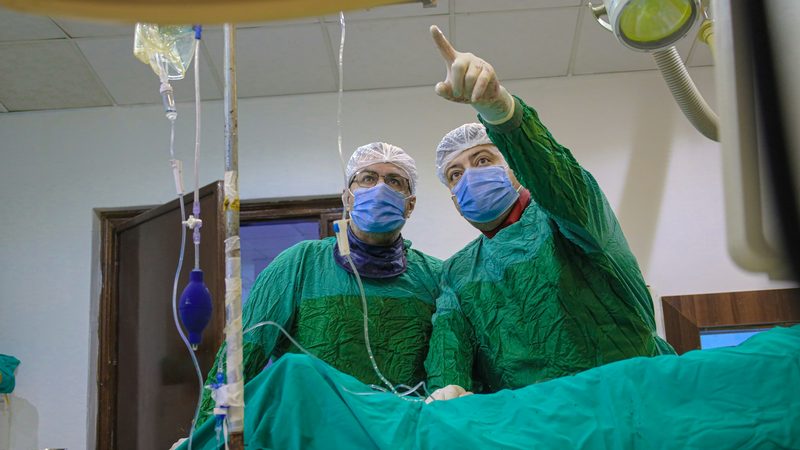 Two clinicians in green gowns, caps and masks stand over a draped patient, one pointing up at a screen out of view
