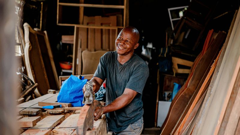A smiling carpenter planes a board in a workshop stacked with timber