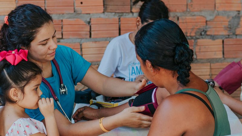 A health worker checks a woman's blood pressure at an outdoor clinic while a girl looks on