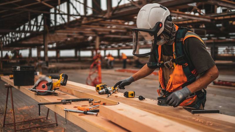 A construction worker in a white hard hat, face shield and orange safety vest lays out tools on a long timber beam under a building frame
