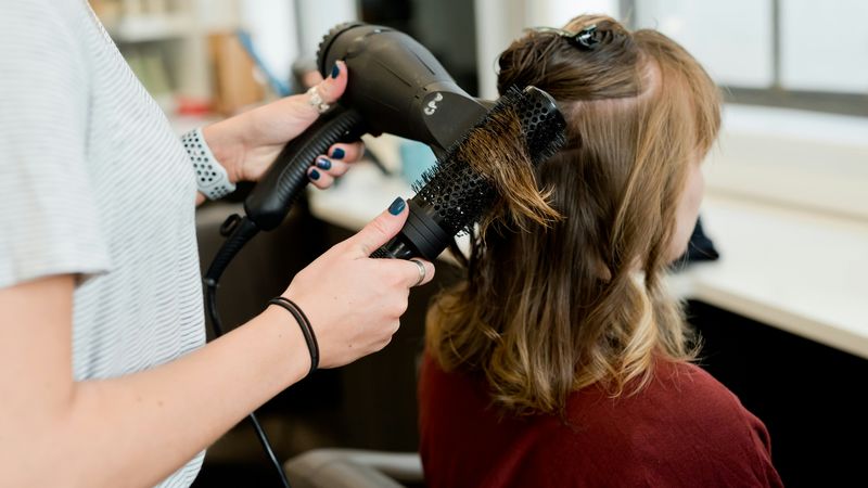 A hairstylist blow-dries a client's hair with a round brush
