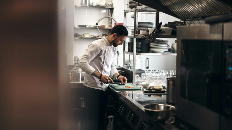 A cook in a white jacket chops vegetables at the counter of a restaurant kitchen