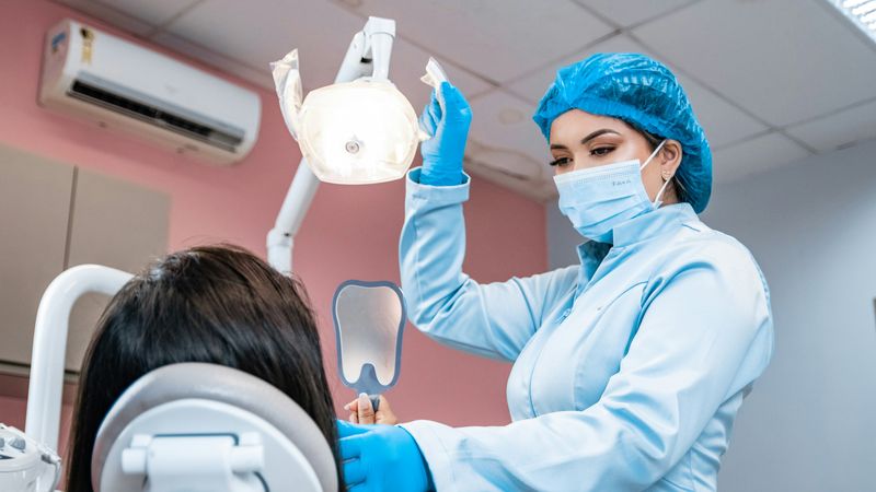 A dental assistant in blue gloves and a hair cover adjusts the light over a patient's chair