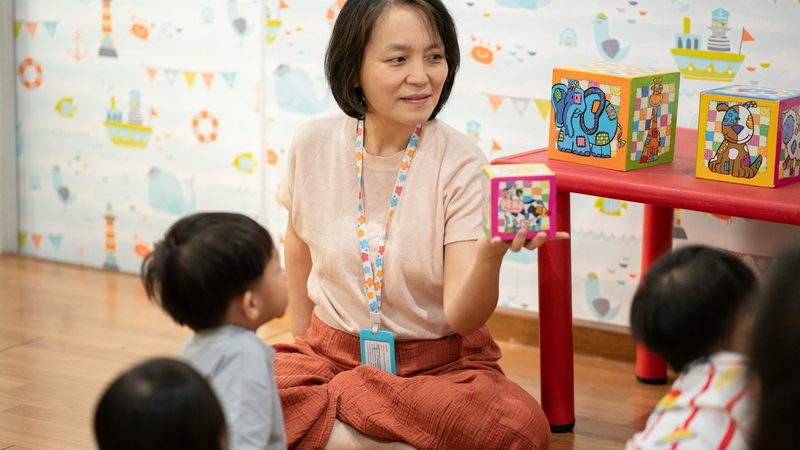 A preschool teacher sits on the floor and shows a picture block to a group of small children