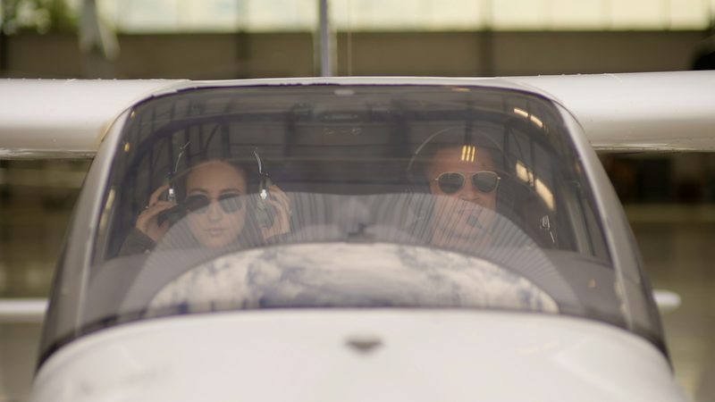 Two pilots in headsets and sunglasses sit behind the windshield of a small white airplane in a hangar
