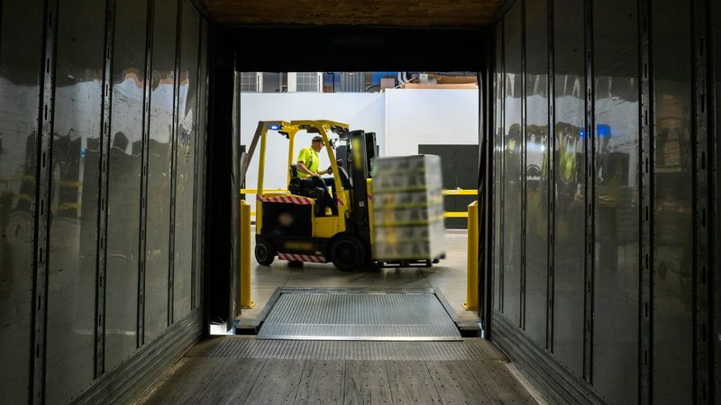 A forklift operator in a safety vest drives a loaded pallet toward the open back of a truck trailer at a loading dock