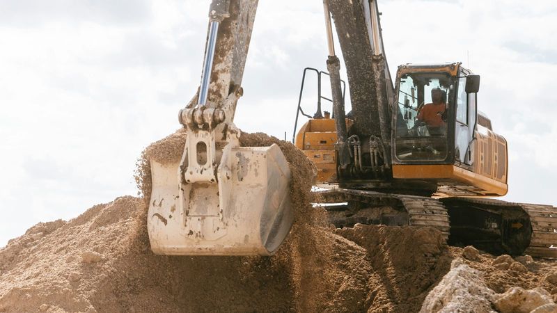 An operator in the cab of an excavator scoops a bucket of earth from a mound