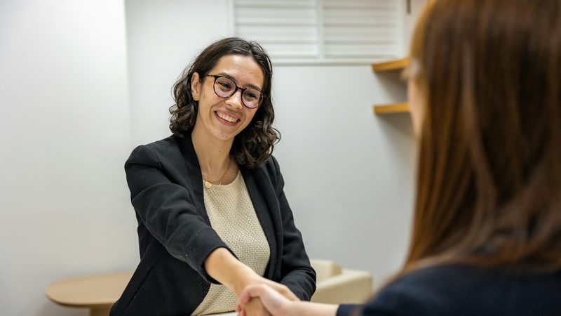 A smiling woman in a blazer shakes hands with a job candidate across a desk