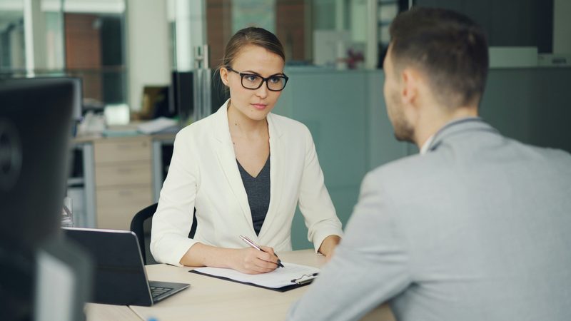 An agent in a white jacket takes notes on a clipboard while talking with a client across her desk