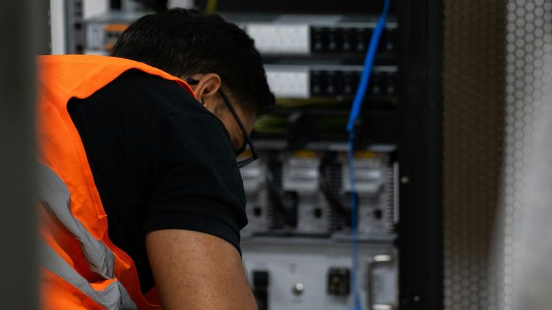 An IT technician in a high-visibility vest works at a rack of network equipment
