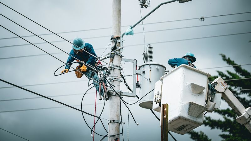 Two line workers in blue hard hats repair wiring on a utility pole, one strapped to the pole and one in a bucket truck