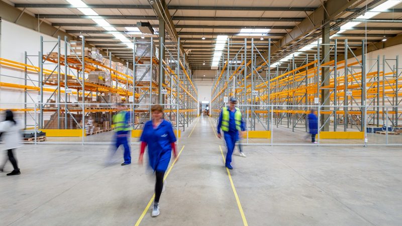 Workers in uniforms and safety vests walk down the aisle of a large warehouse with tall shelving