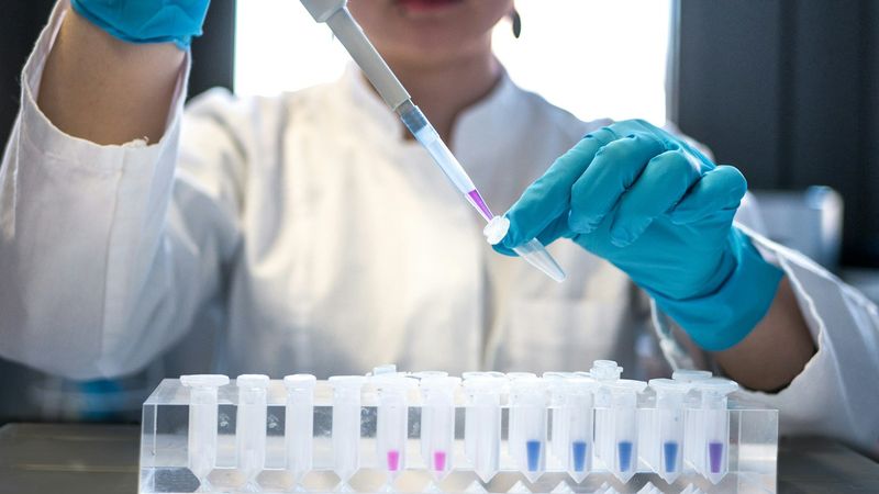 A lab technician in a white coat and blue gloves pipettes a pink sample into a small tube above a rack of test tubes