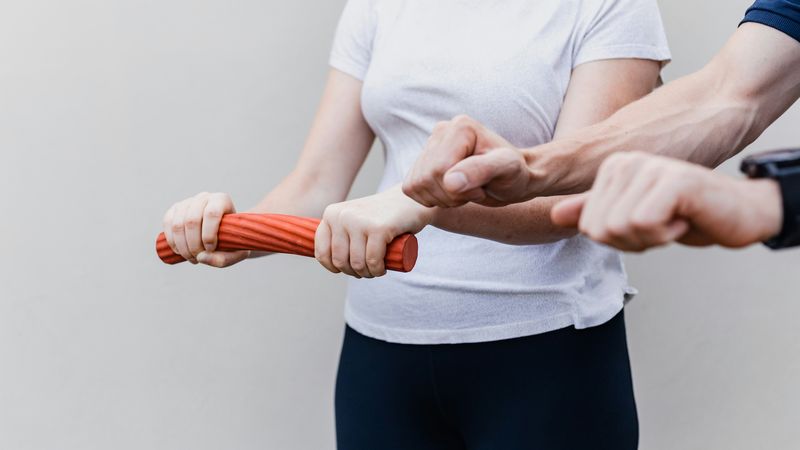 A therapist steadies a woman's wrists as she twists a red therapy bar to build grip strength