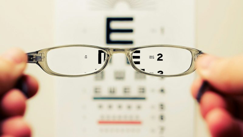 Hands hold up a pair of glasses that bring an eye chart into focus behind them