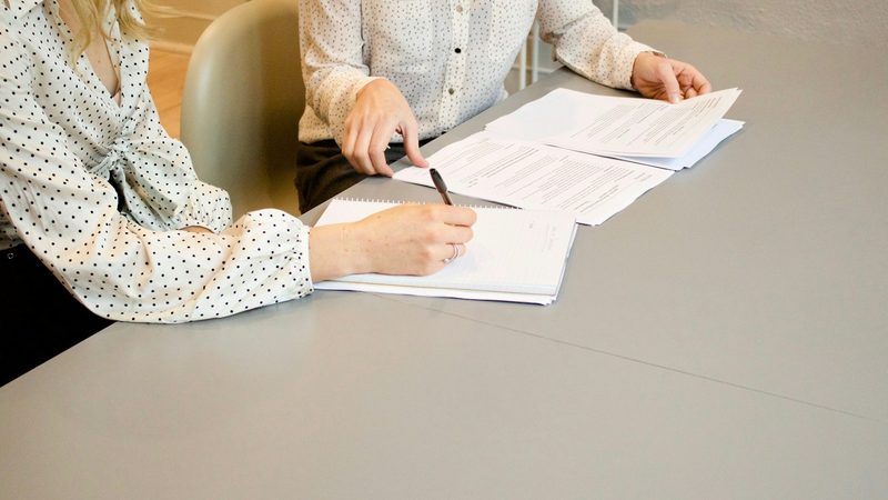 Two women at a desk go through printed documents, one taking notes