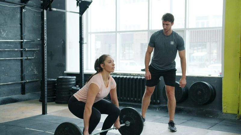 A trainer watches a woman's form as she lifts a barbell from the floor in a gym