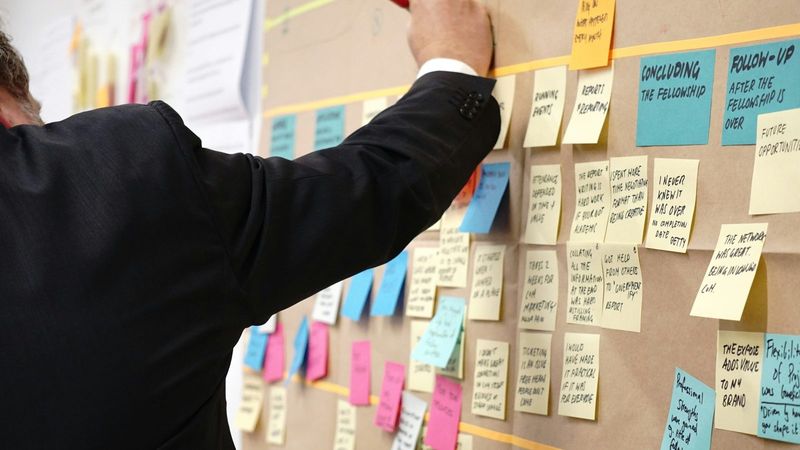 A man in a suit adds a note to a project planning wall covered in sticky notes