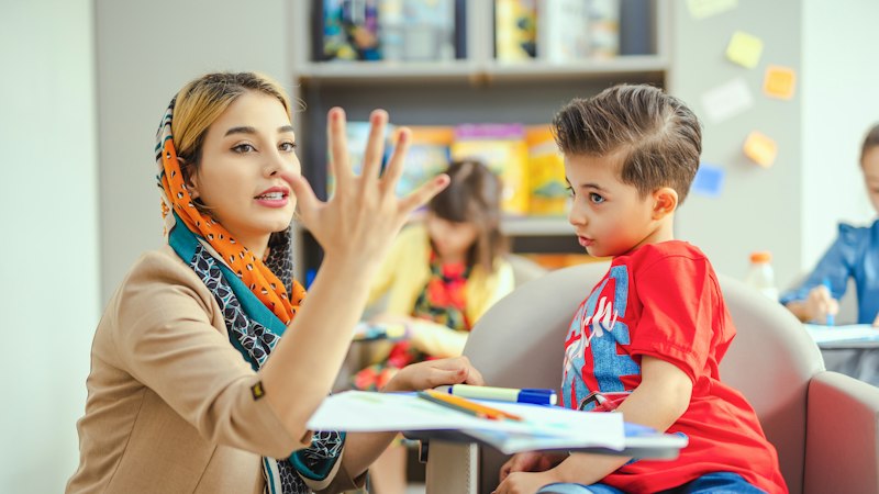 A teacher holds up five fingers for a young boy at a small table in a classroom