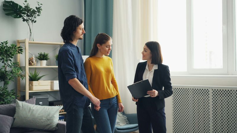 A real estate agent with a clipboard talks with a young couple in the living room of a home