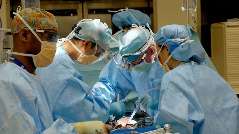 A surgical team in blue gowns, caps and masks at work over an operating table