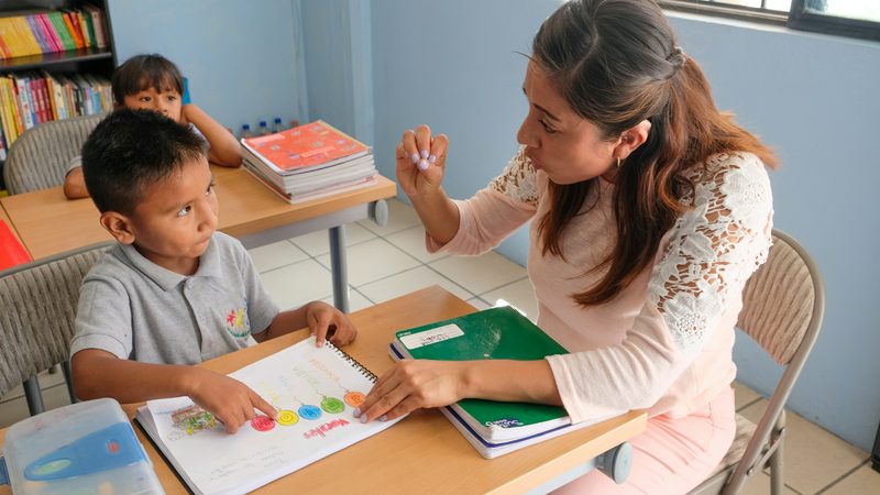 A teacher sits beside a young boy at his desk and holds up a finger while he points to colored circles in his workbook